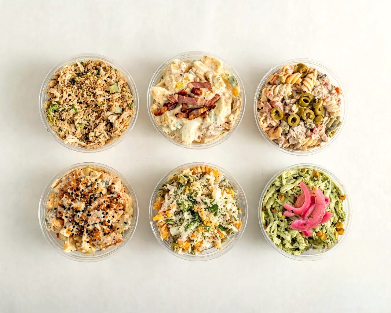 Seis bowls de dels deli vistos desde arriba, en envases para llevar: pollo, pasta, aceitunas, sésamo y cebolla encurtida